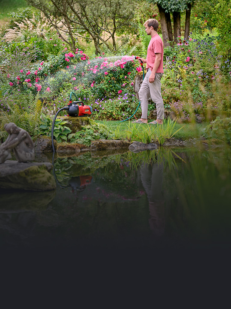 A man waters a red flowering hedge with a hose connected to a red Einhell garden pump.