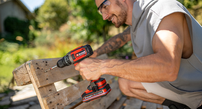 A man wearing safety glasses uses a red and black Einhell cordless drill/driver to work on a wooden bench outdoors.