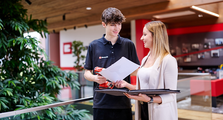 A young employee in an Einhell polo shirt holds a cordless screwdriver and discusses documents with a colleague in a modern office.
