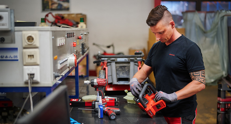 A craftsman in black Einhell clothing assembles or repairs a red Einhell device at a test bench.