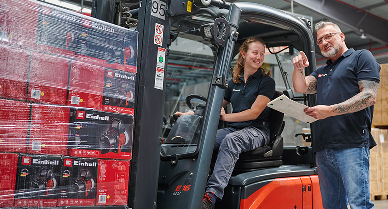 A female employee sits on a forklift next to a pallet of Einhell products and talks to a colleague holding a clipboard.
